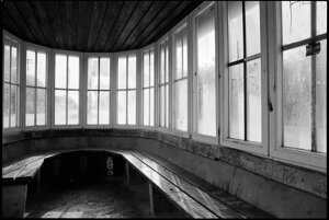 Im Innern des ovalen Pavillons, dem Buswartehäuschens mit den weißen Sprossenfenstern. Blick in Richtung Osten. Hölzerne Sitzbänke im Innern des Buswartehäuschens in Kaköhl. Wer hier Platz nimmt, kann durch die weißen Sprossenfenster buchstäblich in jede Richtung nach draußen sehen, ob wohl gleich der Bus kommt. Diese rund um verlaufende Fensterfront gehört zu den architektonischen Besonderheiten des ovalen Pavillons.